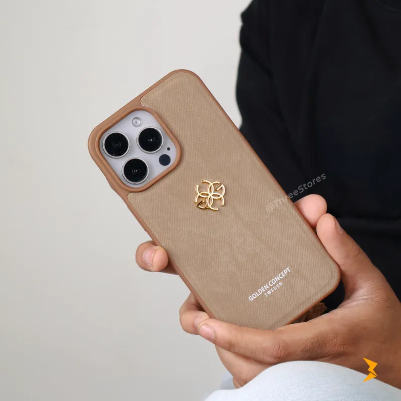 A person holds a tan textured phone case with a gold metal logo and white text on the back, fitted over an iPhone. The background is a clean, neutral off-white wall.
